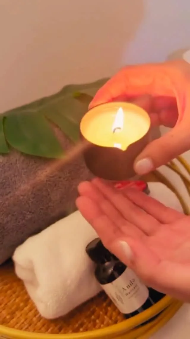 Aromatherapy ritual — hands cradling a lit candle over rolled towels and ANDA oil on a rattan tray